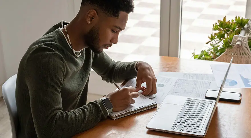 A young man writing notes on a notepad with an open laptop.